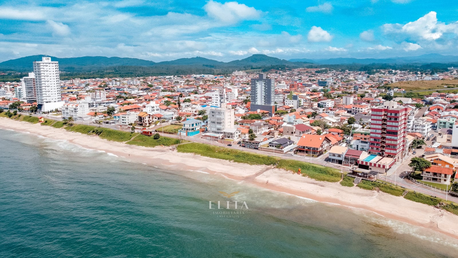 Vista aérea oblíqua de praia com faixa verde e bairro de casas e torres, com morros sob nuvens ao fundo.