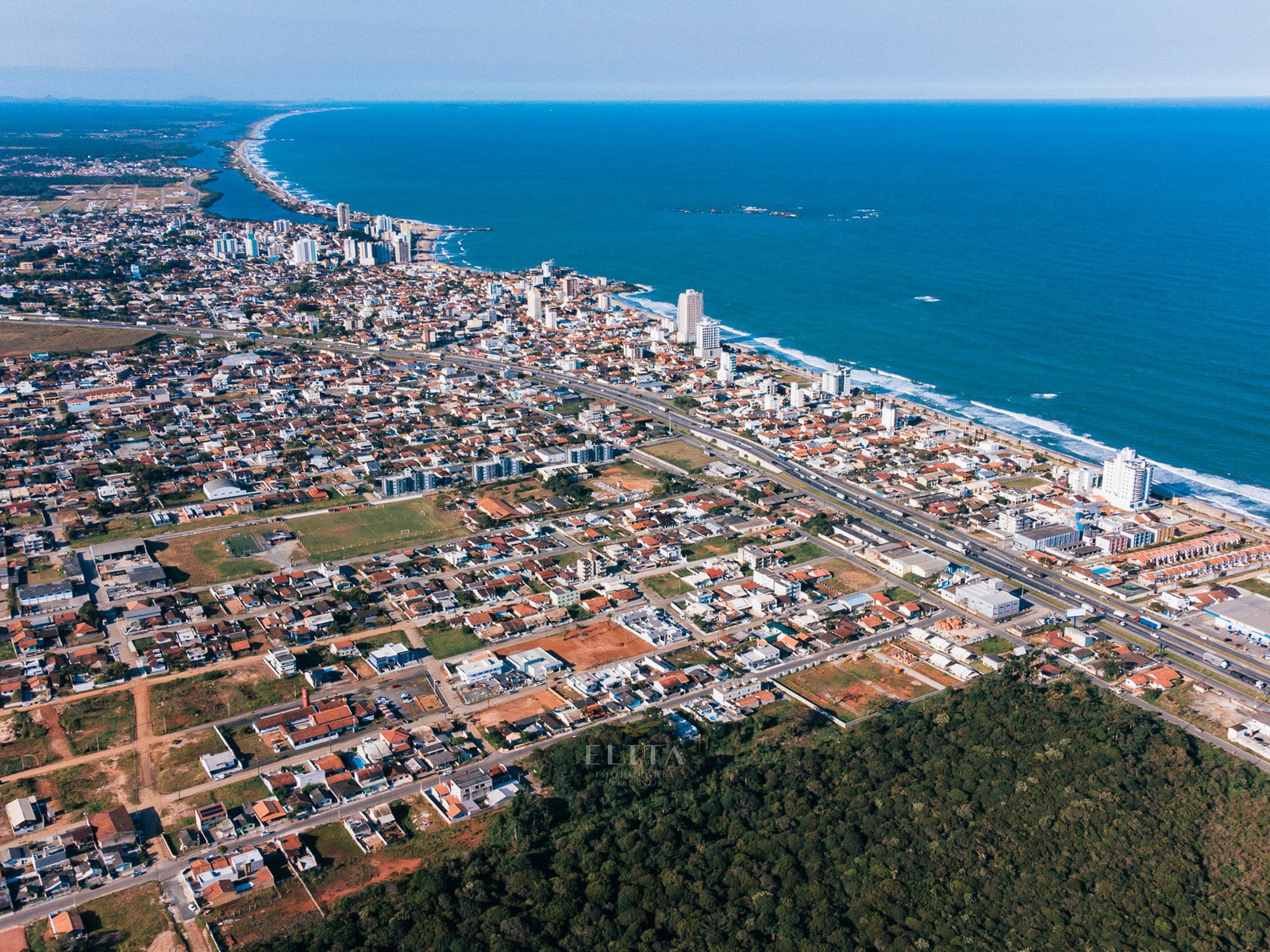 Vista aérea ampla de bairro com casas e terrenos, rodovia de pistas separadas, edifícios na orla e mar ao fundo.