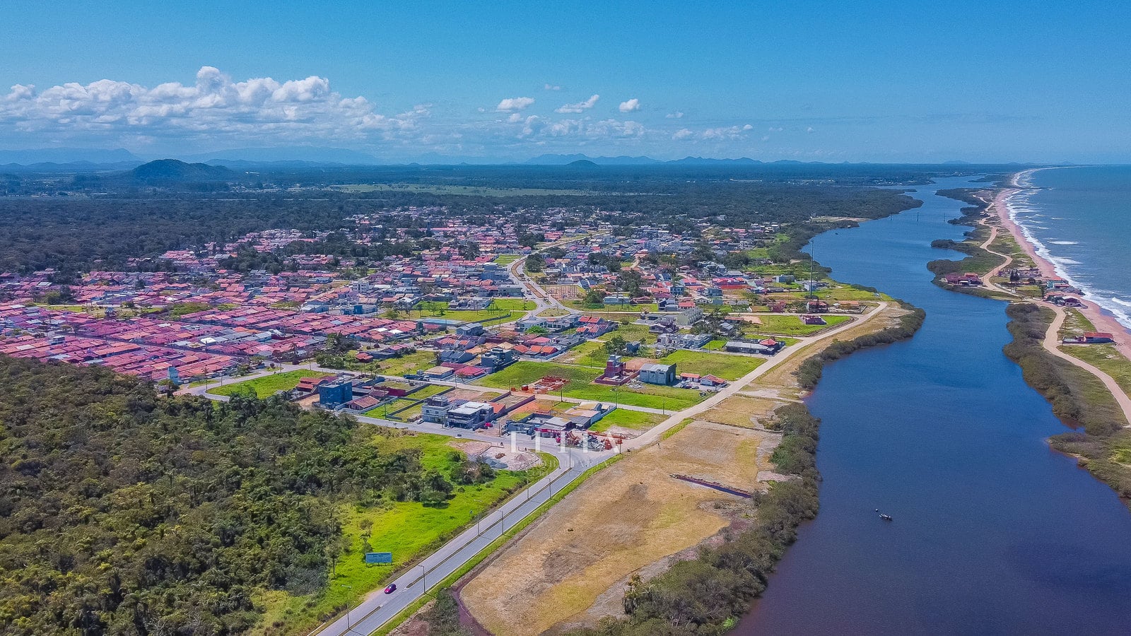 Vista aérea de bairro com casas e terrenos, avenida junto à margem da lagoa e faixa de praia separando a água interior do mar.